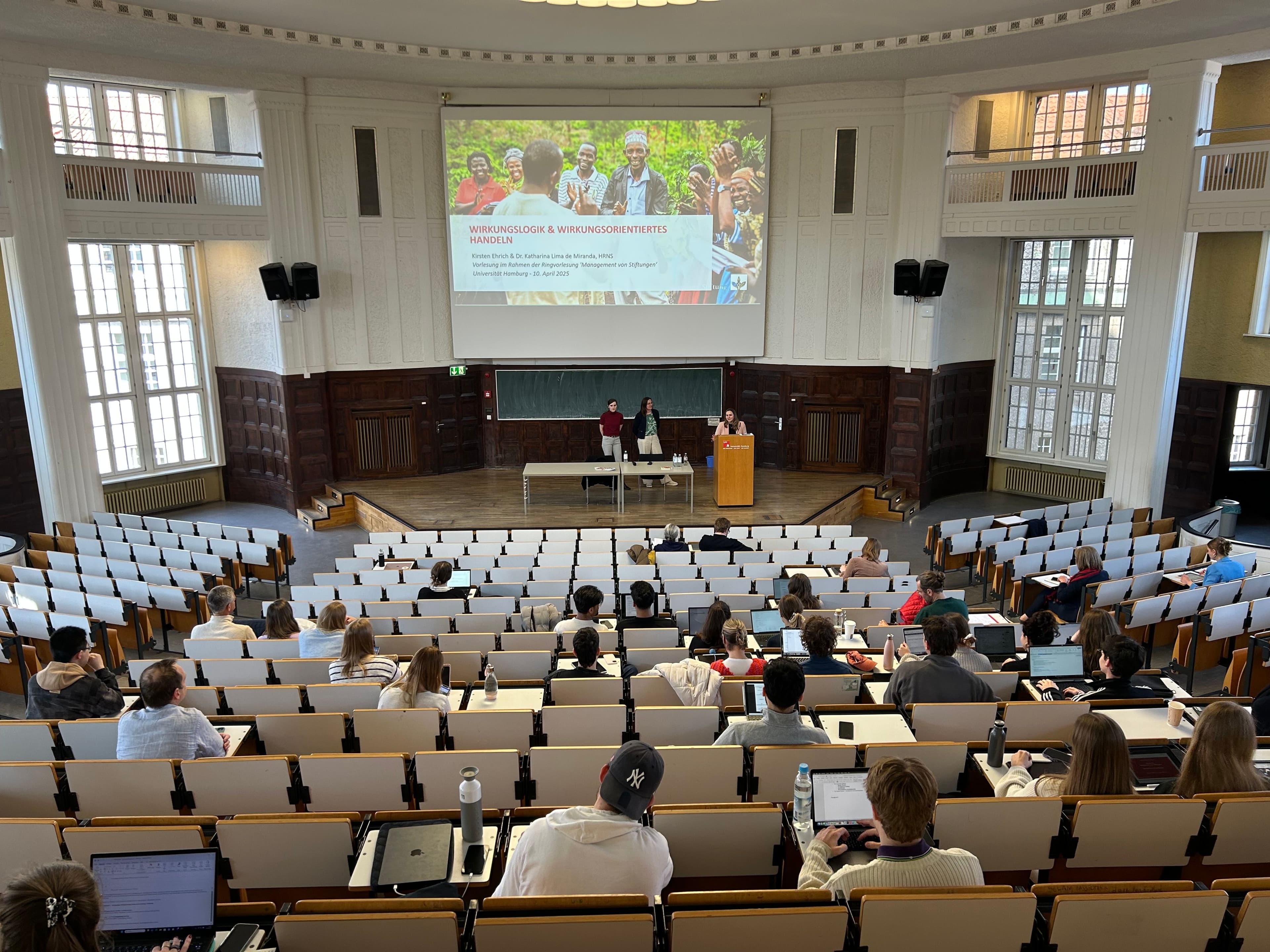 Lecture on Impact Logic and Impact-Oriented Action by HRNS at Universität Hamburg A lecture hall at Universität Hamburg with students seated and a presentation titled "Wirkungslogik & Wirkungsorientiertes Handeln" (Impact Logic & Impact-Oriented Action) projected at the front. Two presenters from HRNS, including Kirsten Ehrich and Dr. Katharina Lima de Miranda, are delivering a talk as part of a course on foundation management.